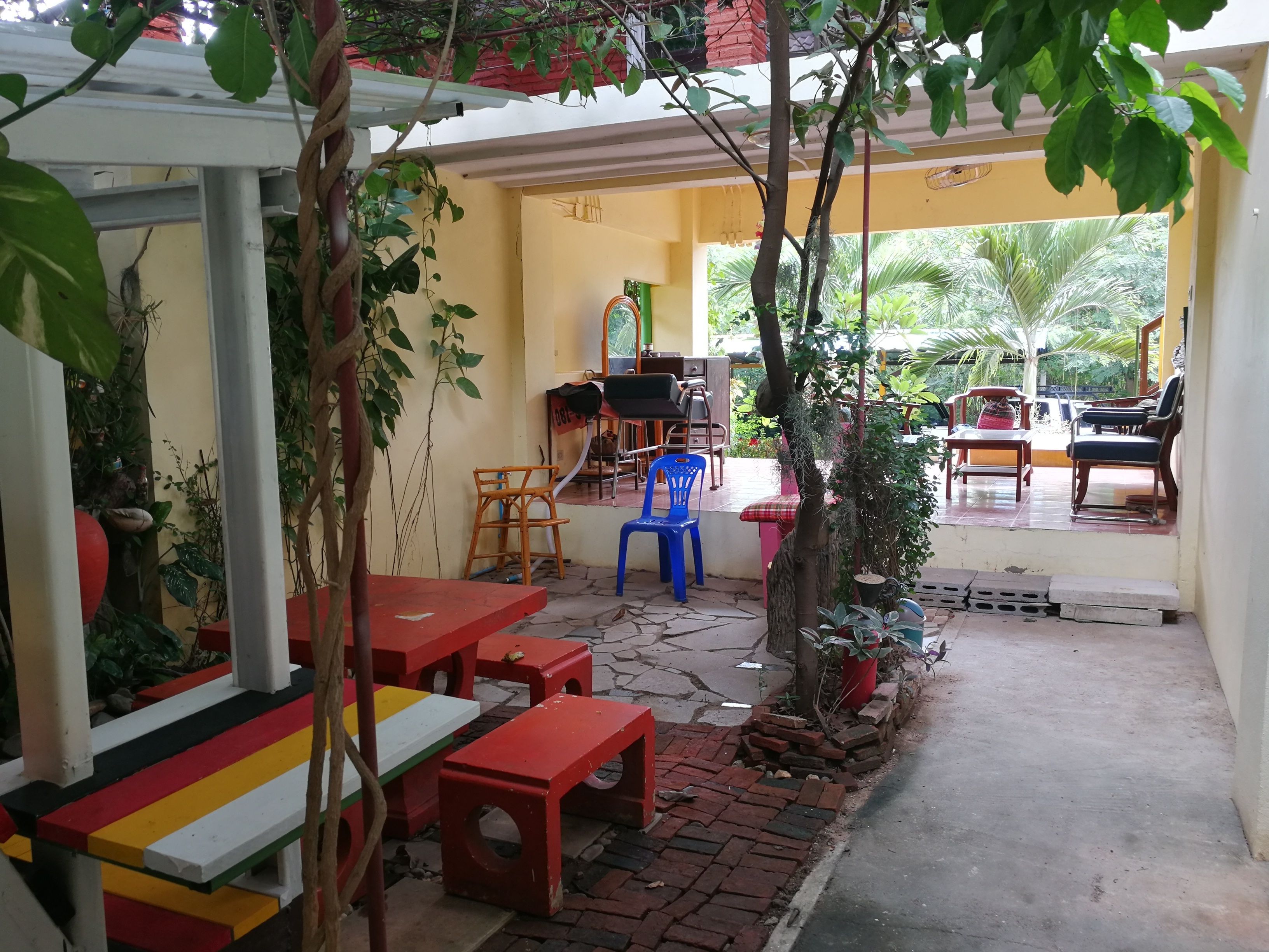 A covered outdoor seating area with tables and chairs, surrounded by plants and trees.