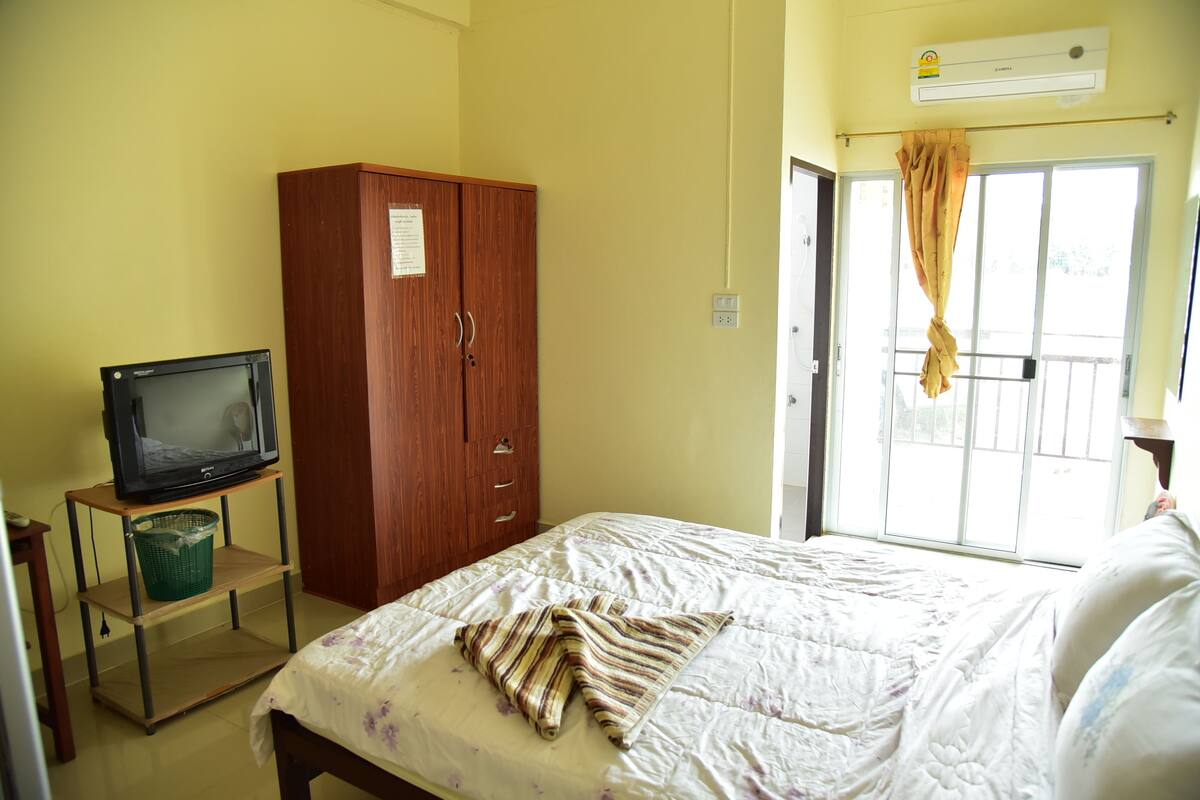Ein Zimmer mit einem Bett, einem Holzschrank, einem Fernseher auf einem Standfuß und einem Fenster mit Vorhang.