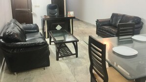 A living room with a sofa, a coffee table, a dining table with chairs, a TV on a stand, and a door. - Jagannath B&B Hotel (New Delhi)