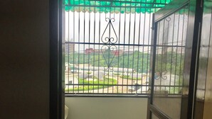 A view through a barred window with a green awning, a metal gate, and a glimpse of a parking area with cars and greenery. - Jagannath B&B Hotel (New Delhi)