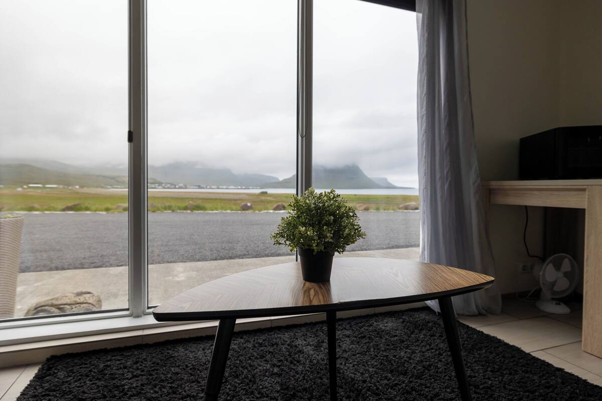 Una habitación con un ventanal grande, una planta en maceta sobre una mesa redonda, un ventilador y vistas a un paisaje lejano.