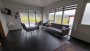 A modern living room with a grey sofa, black chairs, a dining table, and large windows offering a view of the countryside. - Kirkjufell Guesthouse and Apartments (Grundarfjörður)