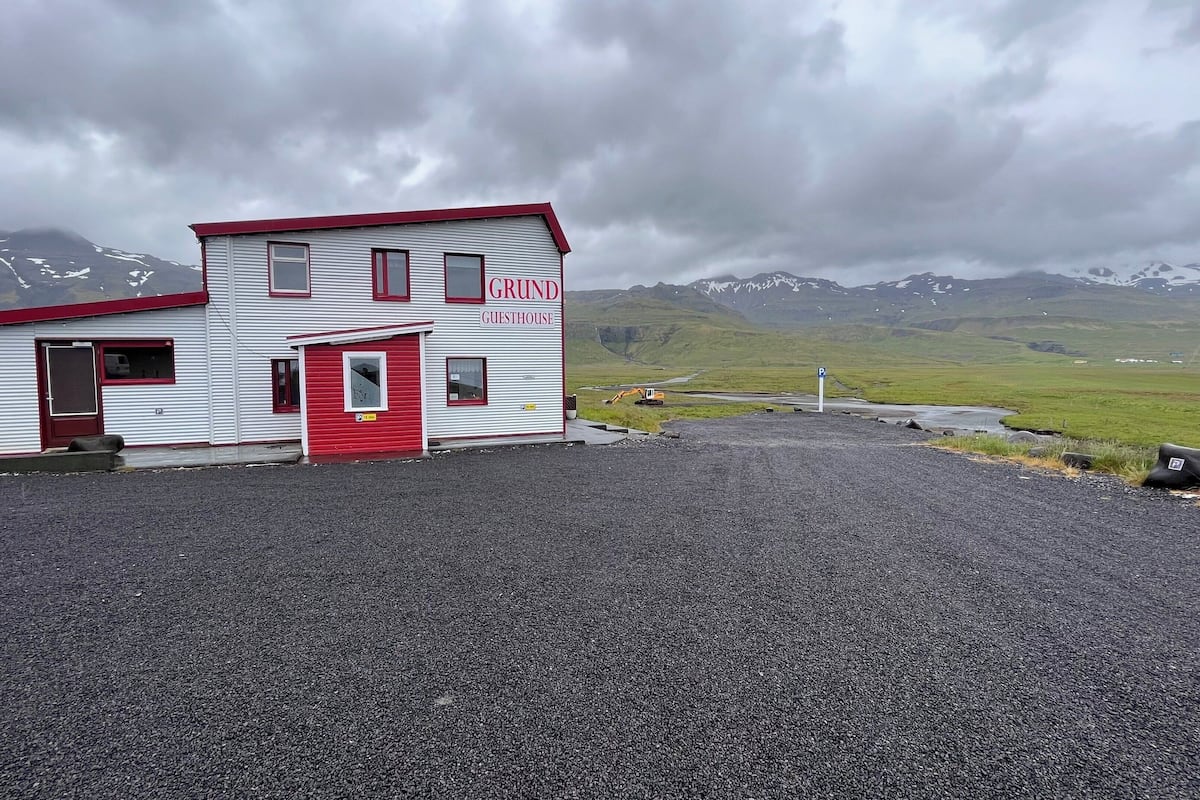 Un bâtiment à deux étages portant l’inscription « GRUND GUESTHOUSE », avec une porte et des fenêtres rouges, situé sur un terrain recouvert de gravier et ayant une chaîne de montagnes en arrière-plan.