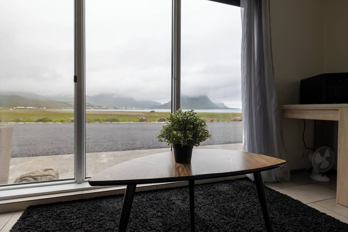 Une pièce dotée d’une grande fenêtre, d’une plante en pot posée sur une table ronde, d’un ventilateur et offrant une vue sur un paysage lointain.