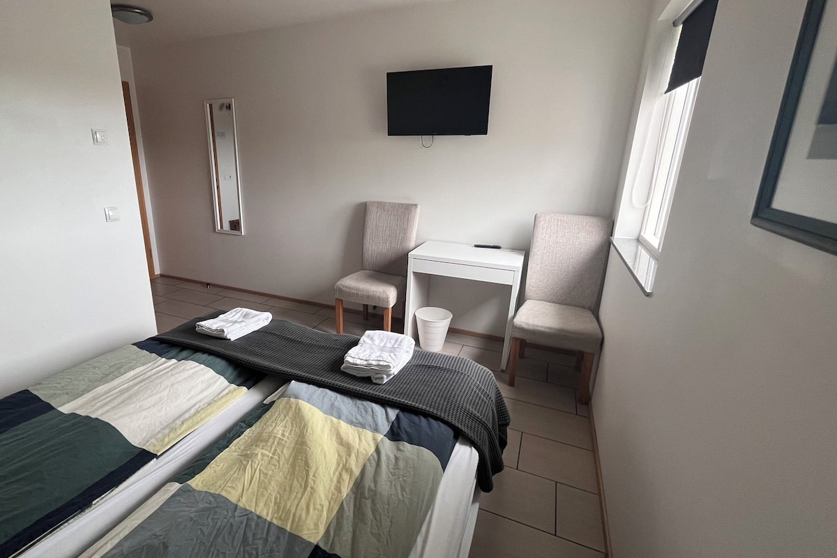 Une petite chambre propre, avec deux lits simples, un bureau, une chaise, une télévision et une fenêtre.