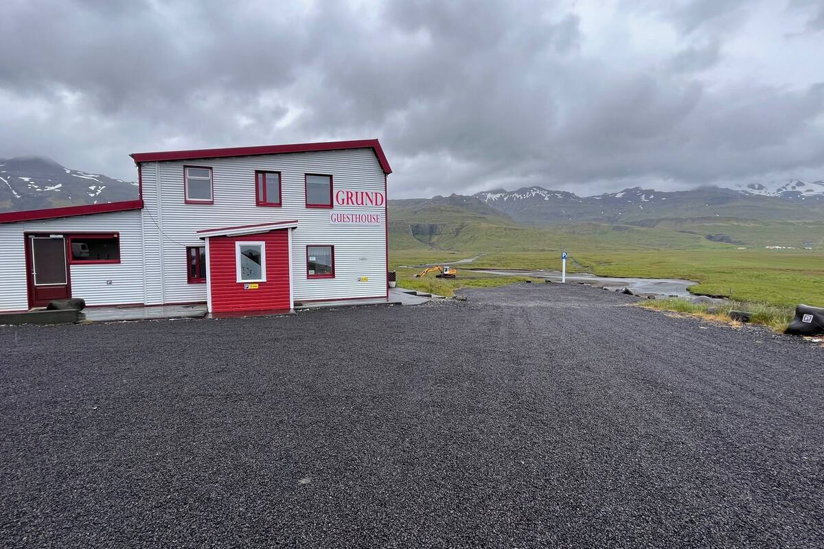 Un edificio de dos pisos con un letrero que dice "GRUND GUESTHOUSE", una puerta roja y ventanas, ubicado en un estacionamiento de grava con montañas al fondo.