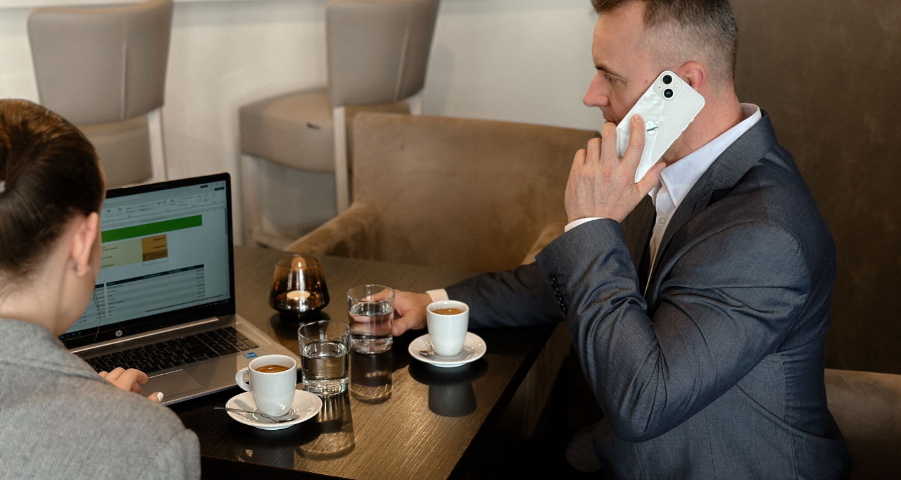 Un hombre con traje habla por teléfono en una mesa, junto a un ordenador portátil, dos tazas de café y vasos de agua.