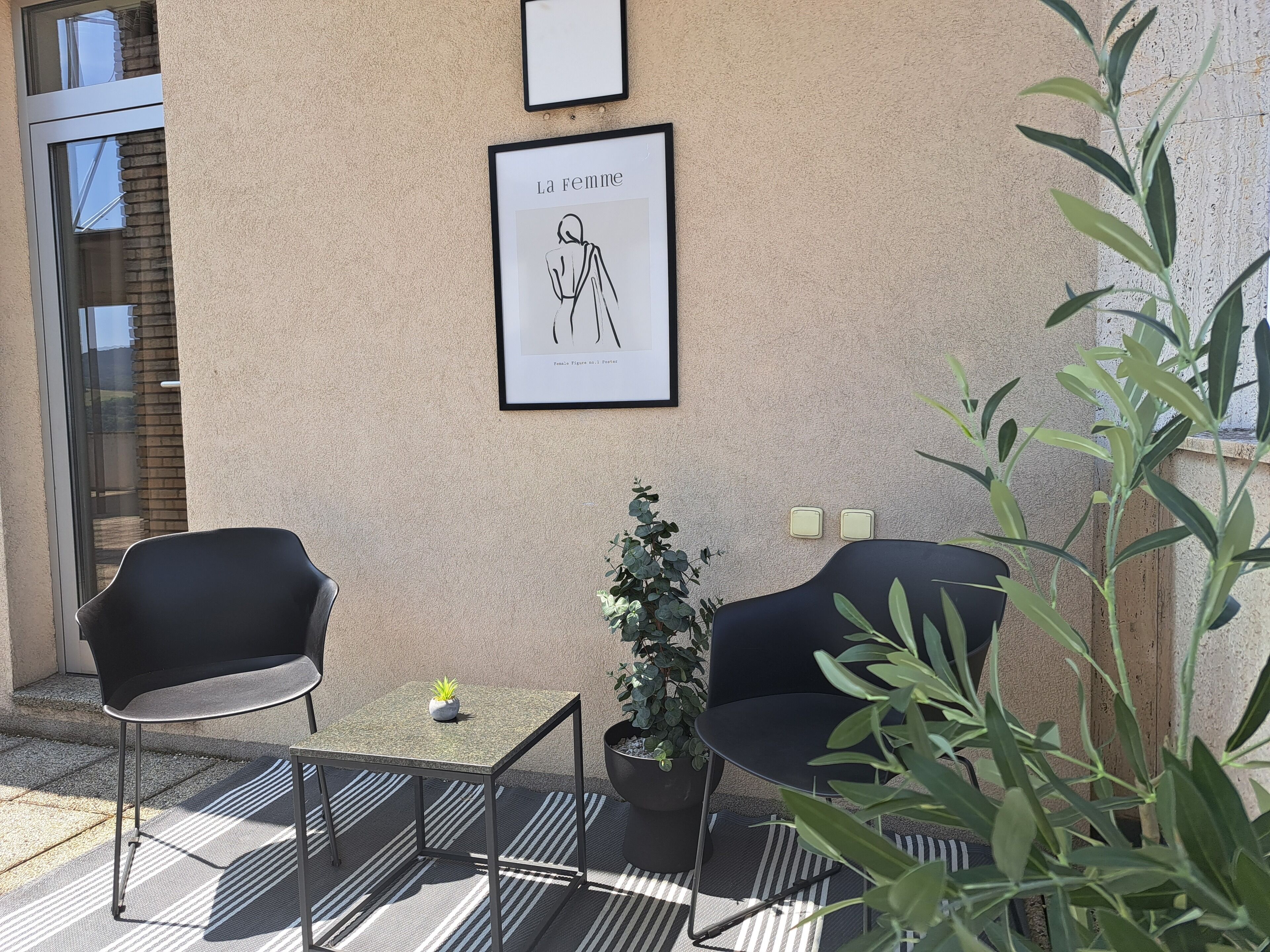 A balcony with two black chairs, a small table, and a potted plant. There is a framed artwork above the table.
