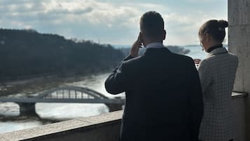 Twee personen staan bij een rivier, met een brug op de achtergrond.