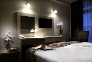 Habitación de hotel con cama, mesita de noche, lámpara, jarrón con flores y televisor de pantalla plana montado en la pared.