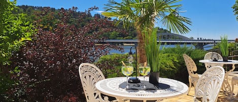 An outdoor dining area with white metal chairs and tables, a palm tree, and a view of a bridge over water.