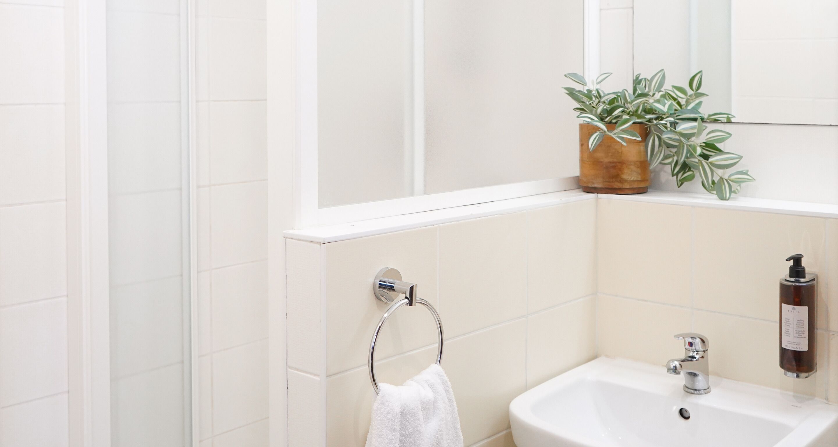 Baño con azulejos blancos, lavamanos, espejo, toallero y planta en maceta.