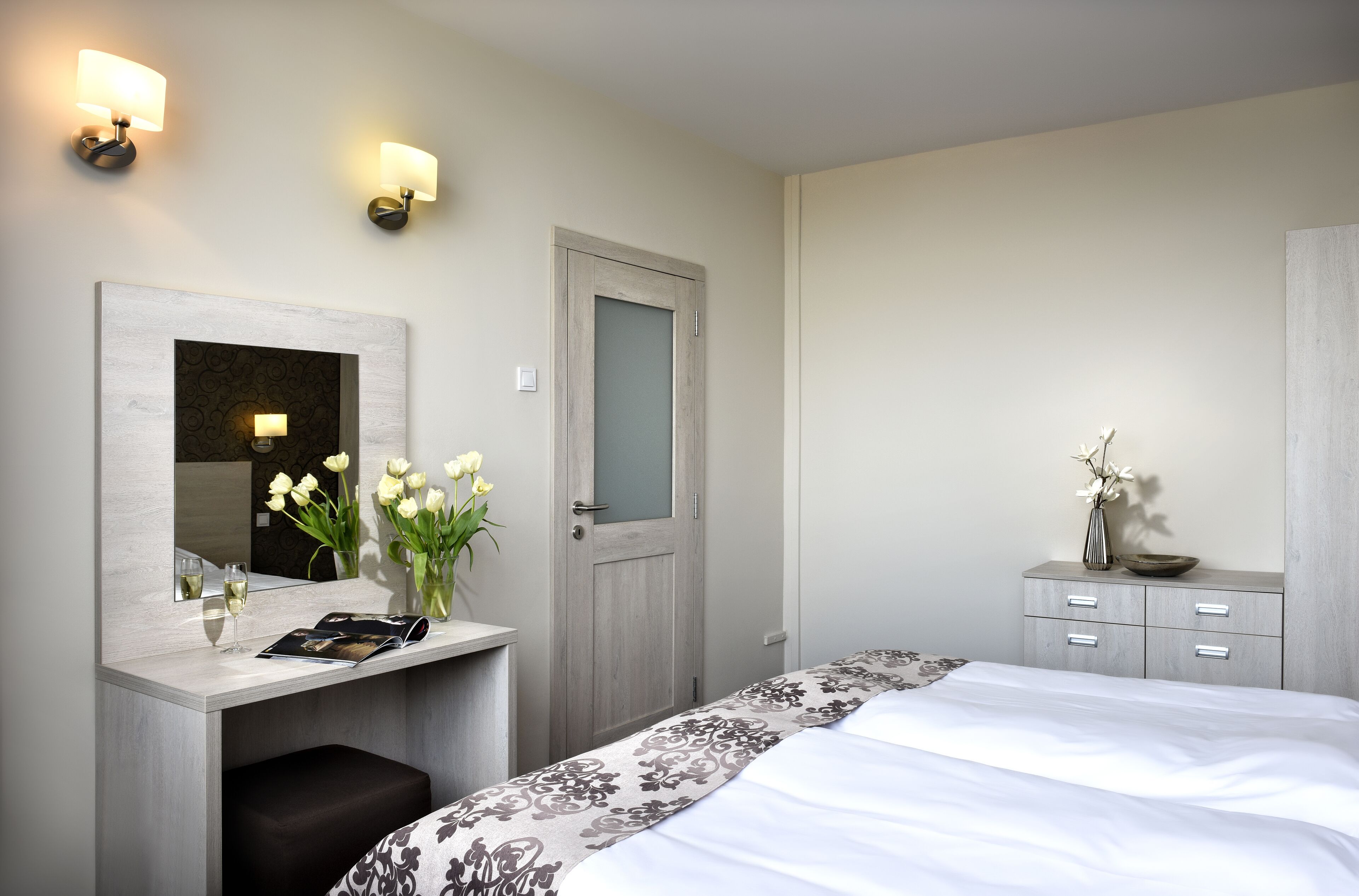 Habitación de hotel con cama, mesita de noche, espejo y un pequeño escritorio con un jarrón de flores y una revista.