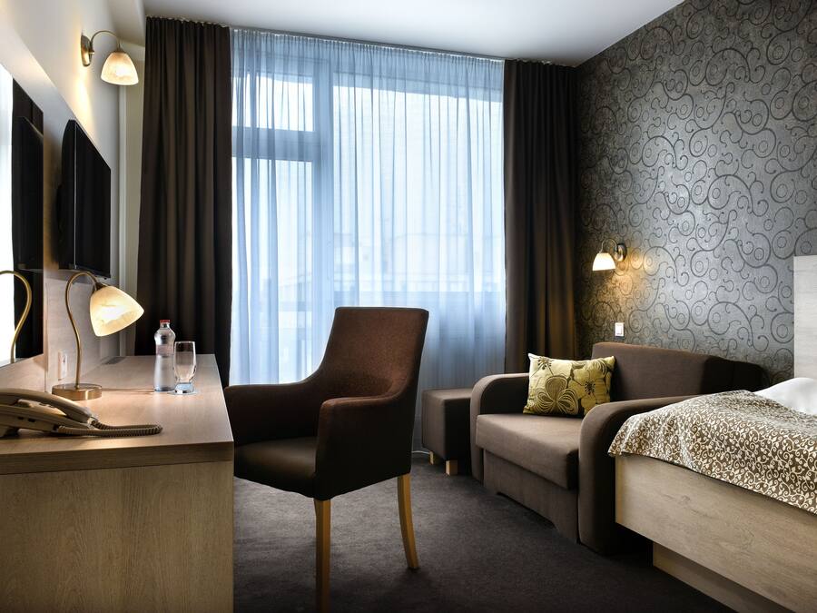 Ein Hotelzimmer mit Schreibtisch, Stuhl, Sofa und Bett. Es gibt zwei Nachttischlampen, einen Spiegel und ein Fenster mit Vorhängen.