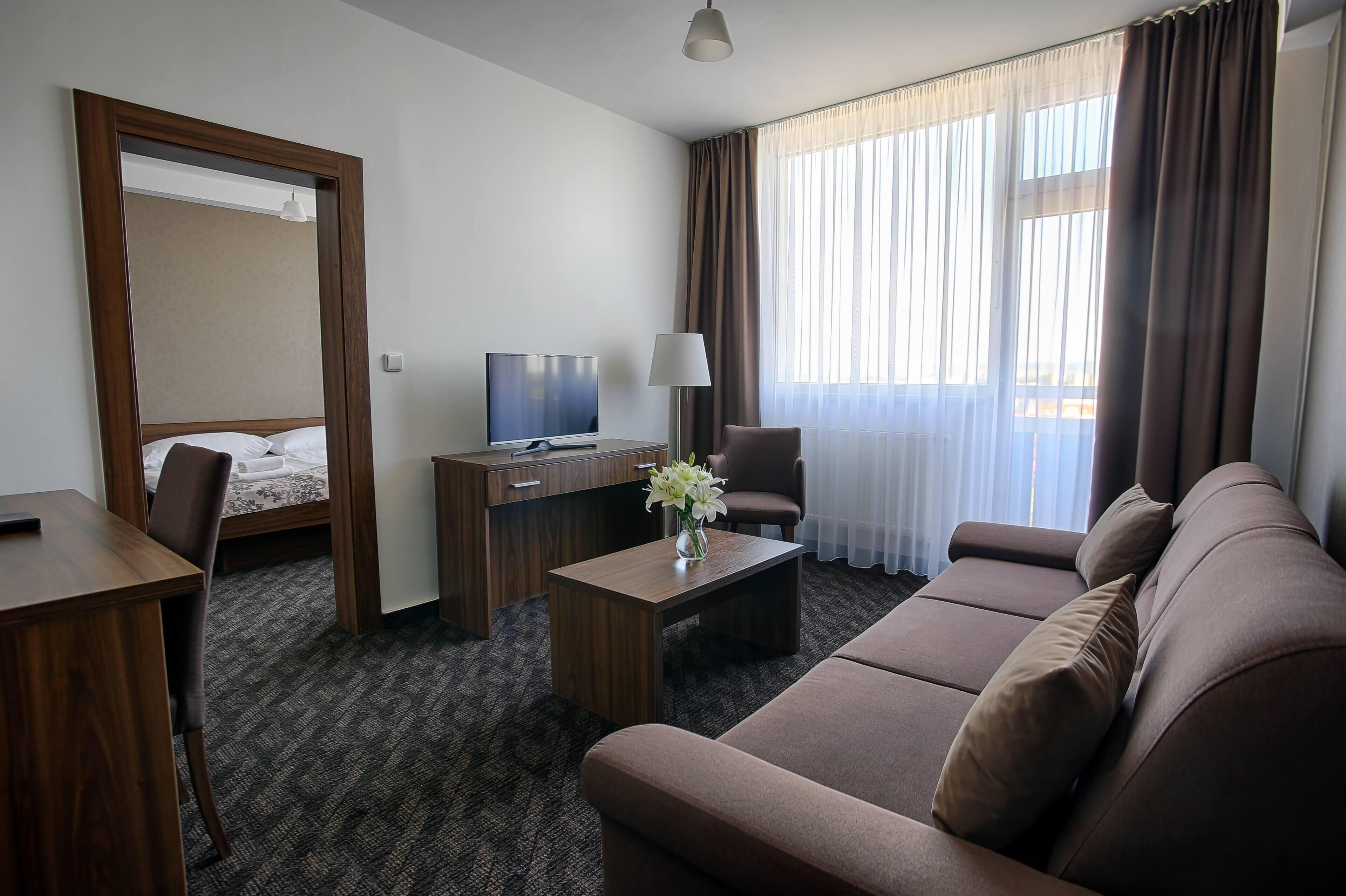 A hotel room with a bed, a desk with a TV, a sofa, a coffee table with a vase, and a large window with curtains.