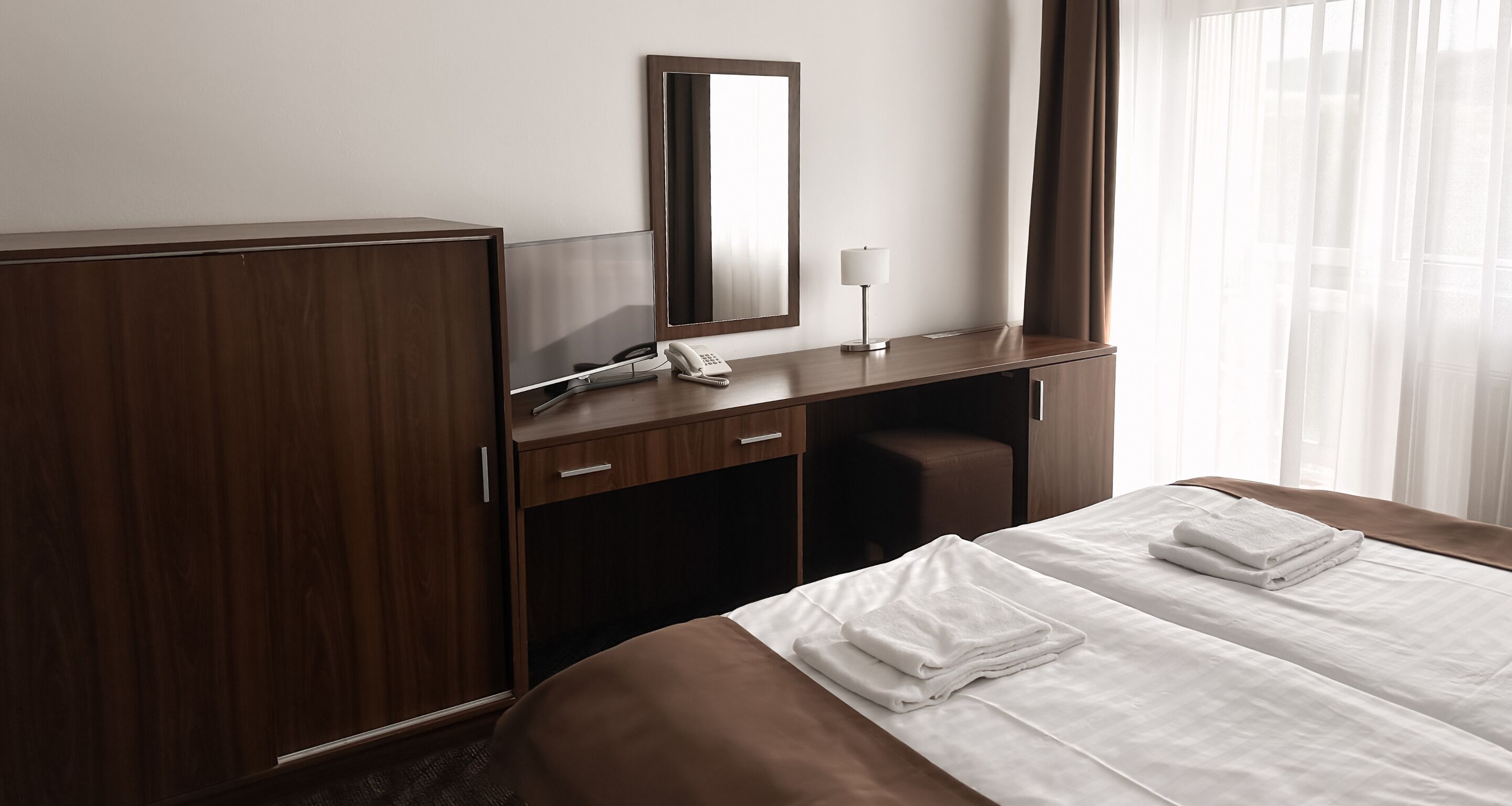A hotel room with a large bed, a desk with a telephone, a mirror, and a window with curtains.