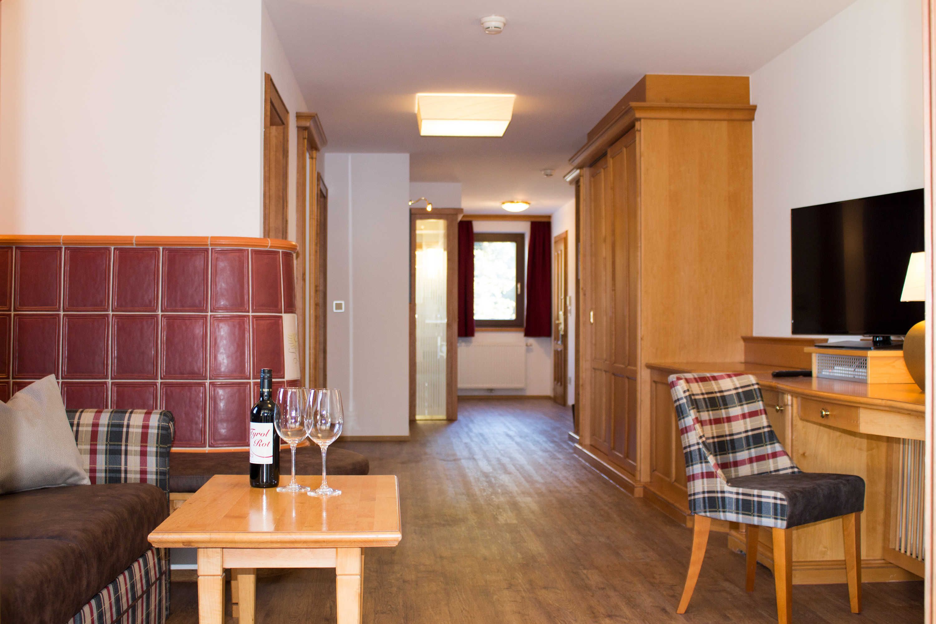 Quarto de hotel com sofá, uma mesa de madeira com uma garrafa e copos, uma poltrona xadrez, televisão e um guarda-roupa de madeira.