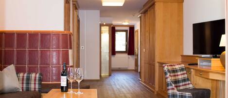 Quarto de hotel com sofá, uma mesa de madeira com uma garrafa e copos, uma poltrona xadrez, televisão e um guarda-roupa de madeira.