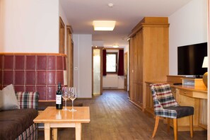 A hotel room with a sofa, a wooden table with a bottle and glasses, a plaid chair, a TV, and a wooden wardrobe. - Comfort Suite TYROL - Hotel Tyrol am Haldensee (Haldensee)