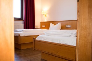 A hotel room with two wooden beds, a window with red curtains, a wall-mounted lamp, and a radiator. - Comfort Suite TYROL - Hotel Tyrol am Haldensee (Haldensee)
