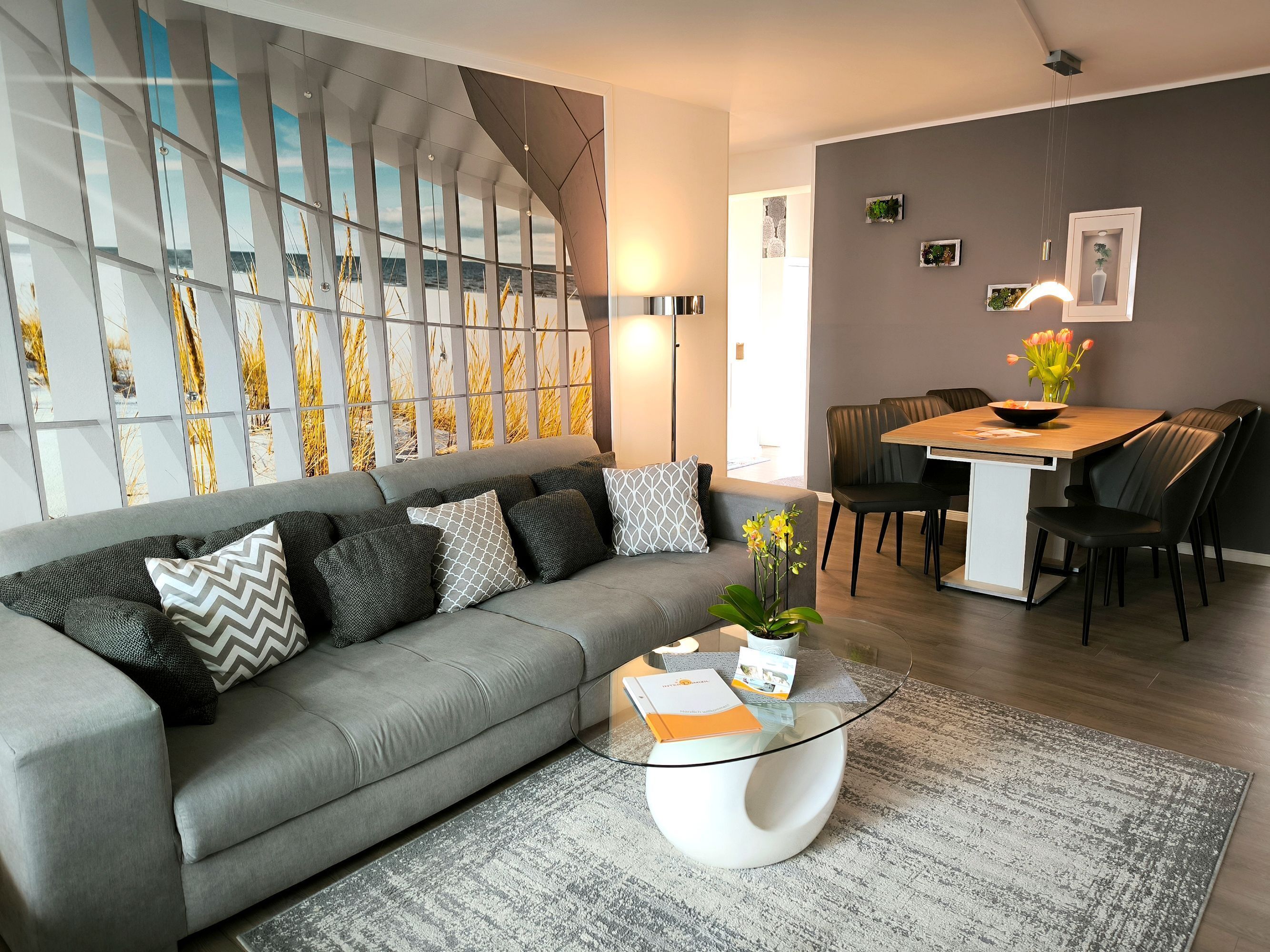 Una sala de estar moderna con un sofá gris, una mesa de centro de vidrio y una zona de comedor con una mesa y sillas de madera.