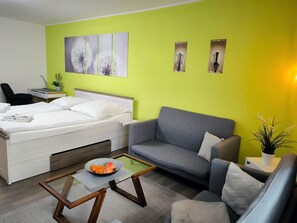 Quarto de hotel moderno com uma cama, um sofá cinzento, uma mesa de centro de vidro e uma pequena mesa lateral com uma planta.