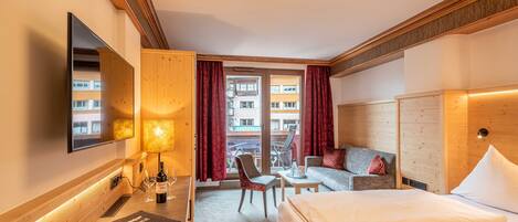 Quarto de hotel com cama, sofá, escrivaninha com abajur, cadeira, televisão e janela com cortinas.