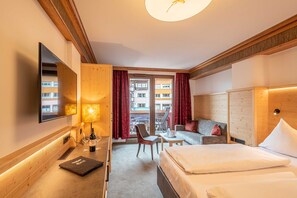 Habitación de hotel con cama, sofá, escritorio con lámpara, silla, televisor y ventana con cortinas.