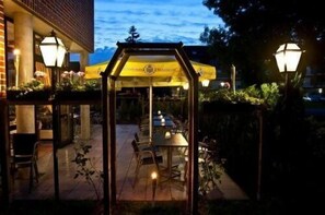 An outdoor patio with a covered seating area and tables, surrounded by greenery and a brick building.