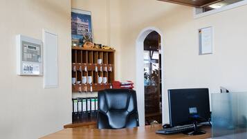 A reception desk with a computer, a black office chair, and a glass partition. There is a wooden shelf with various items and a poster on the wall.