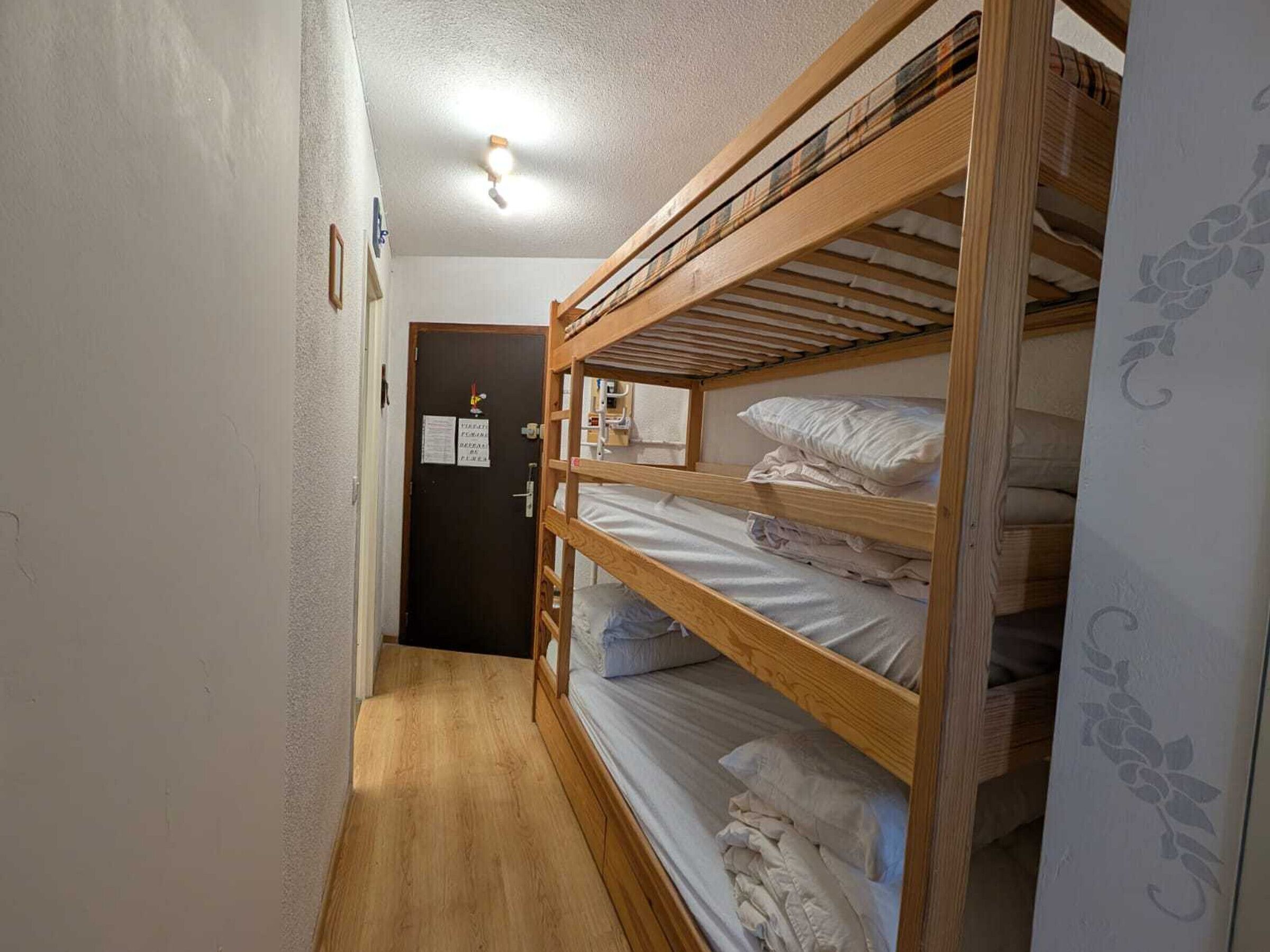 A narrow hallway with wooden bunk beds, a door with a notice, and a light fixture on the ceiling.