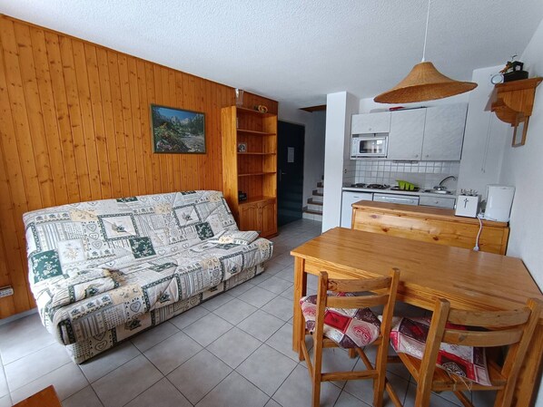 Ein gemütliches Wohnzimmer mit einem gemusterten Sofa, einem Holzesstisch und Stühlen. Im Hintergrund ist eine Küche mit weißen Schränken und einer Hängelampe zu sehen.
