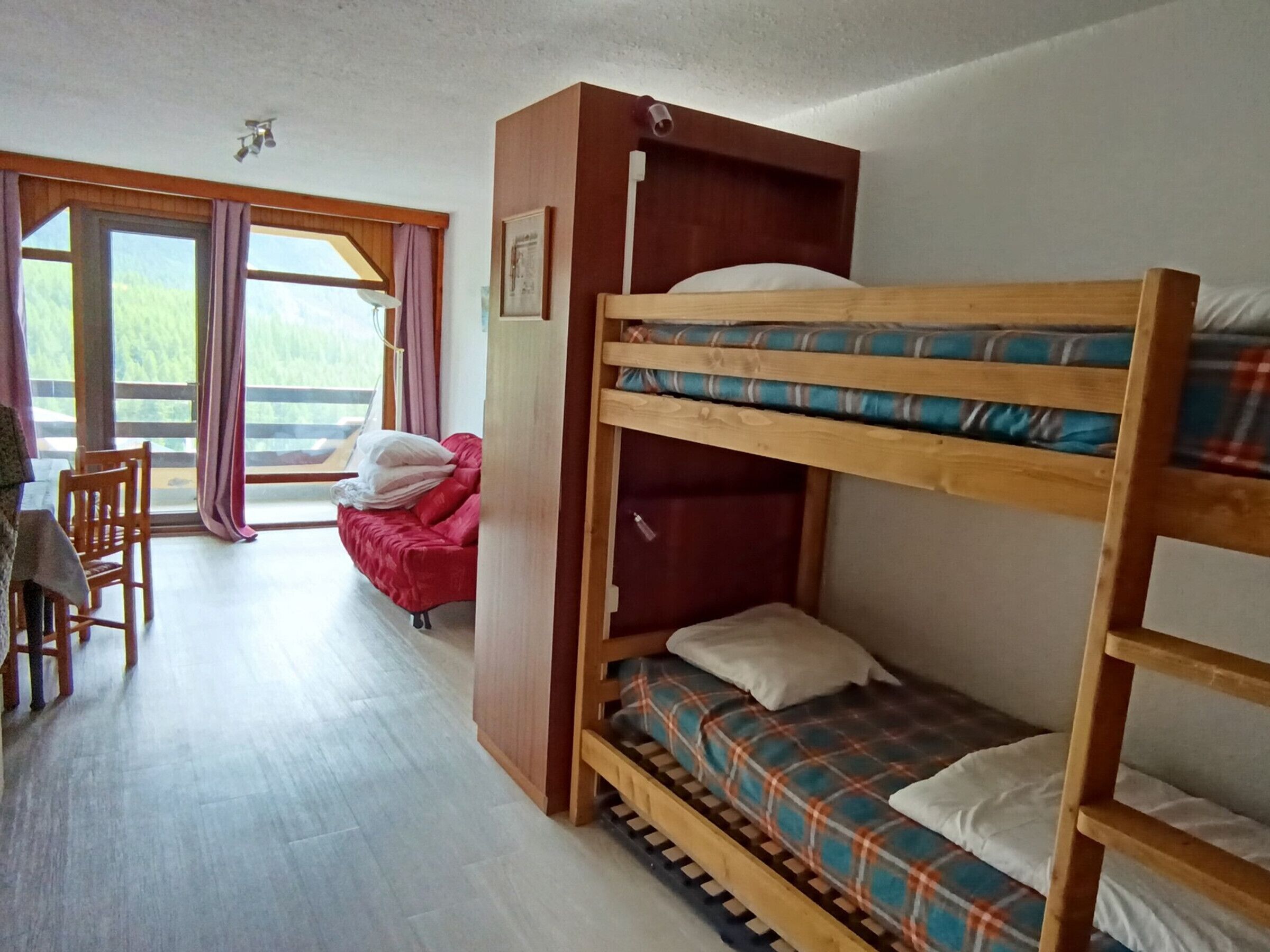 A dormitory room with bunk beds, a red sofa, a wooden desk, and a large window with curtains.
