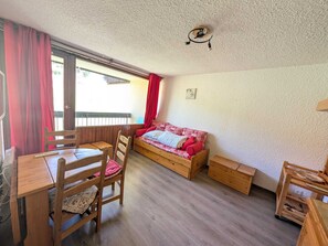 A room with a wooden table, chairs, a bed with red bedding, and a small wooden cabinet.