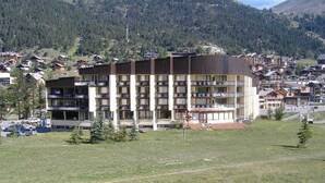 A multi-story hotel with a flat roof and balconies, surrounded by a grassy field and a mountainous backdrop.
