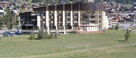A multi-story hotel with a flat roof and balconies, surrounded by a grassy field and a mountainous backdrop.