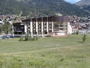 A multi-story hotel with a flat roof and balconies, surrounded by a grassy field and a mountainous backdrop.