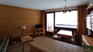 A room with wooden walls, a tiled floor, a bed, a desk, a chair, a table, a TV, and a window with curtains. - Spacious apartment on slopes, sleeps 4-6, nice view (Montgenèvre)