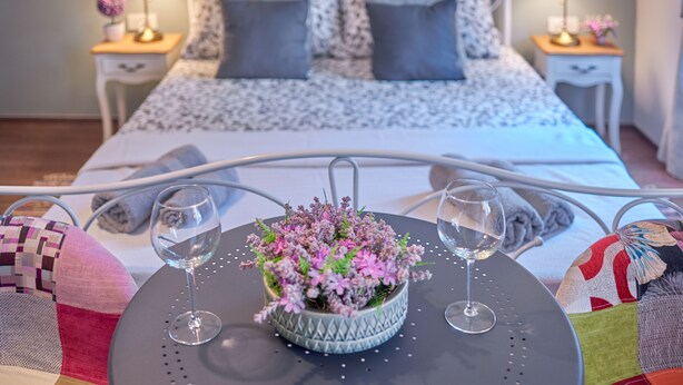 Una cama bien hecha con una colcha estampada, dos lámparas de noche y una mesita con una planta en maceta y dos copas de vino.