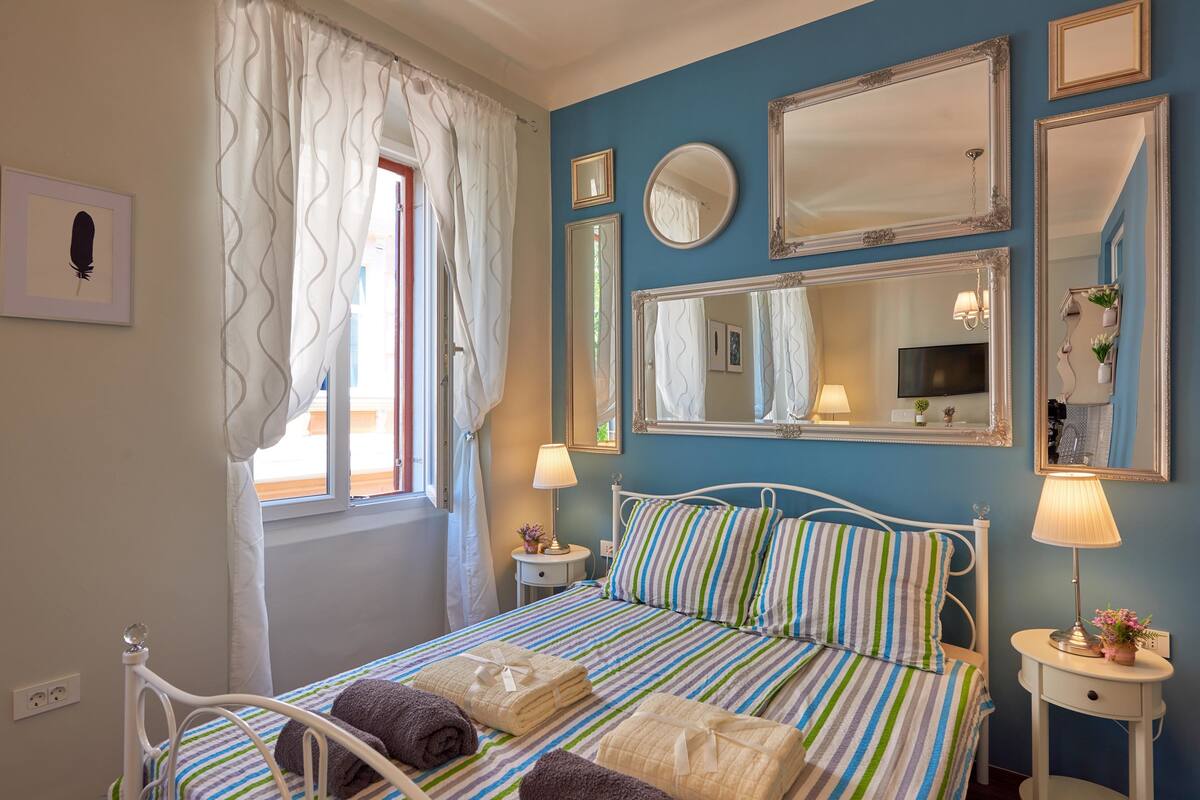 A bedroom with a bed, bedside tables, a window with curtains, and wall-mounted mirrors.