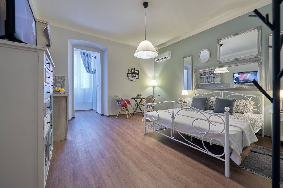 A bedroom with a bed, bedside tables, a desk, and a chair. There is a window with curtains, a hanging light fixture, and a mirror on the wall.