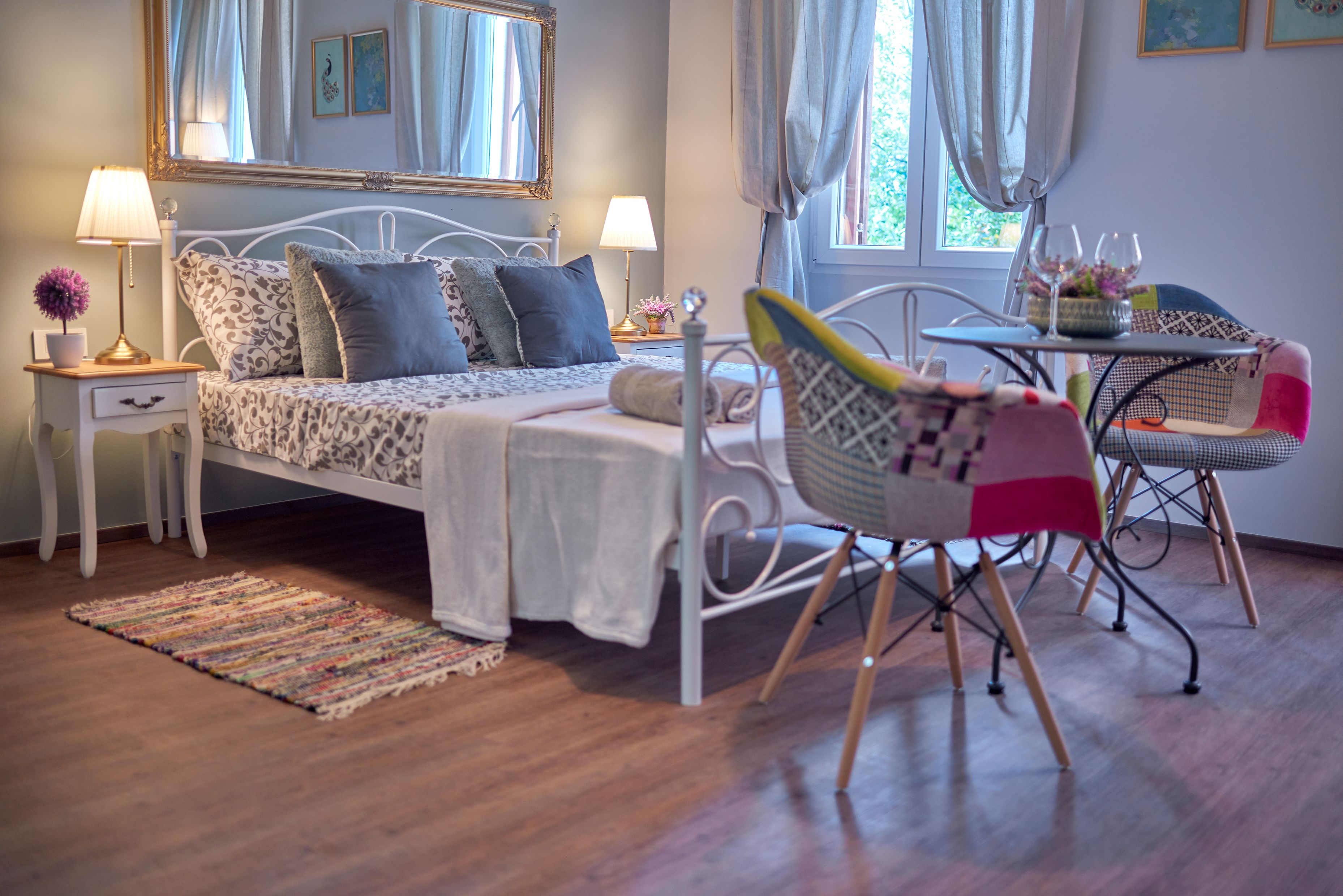 Un dormitorio con cama, mesitas de noche, una silla, una mesita pequeña y una ventana con cortinas.