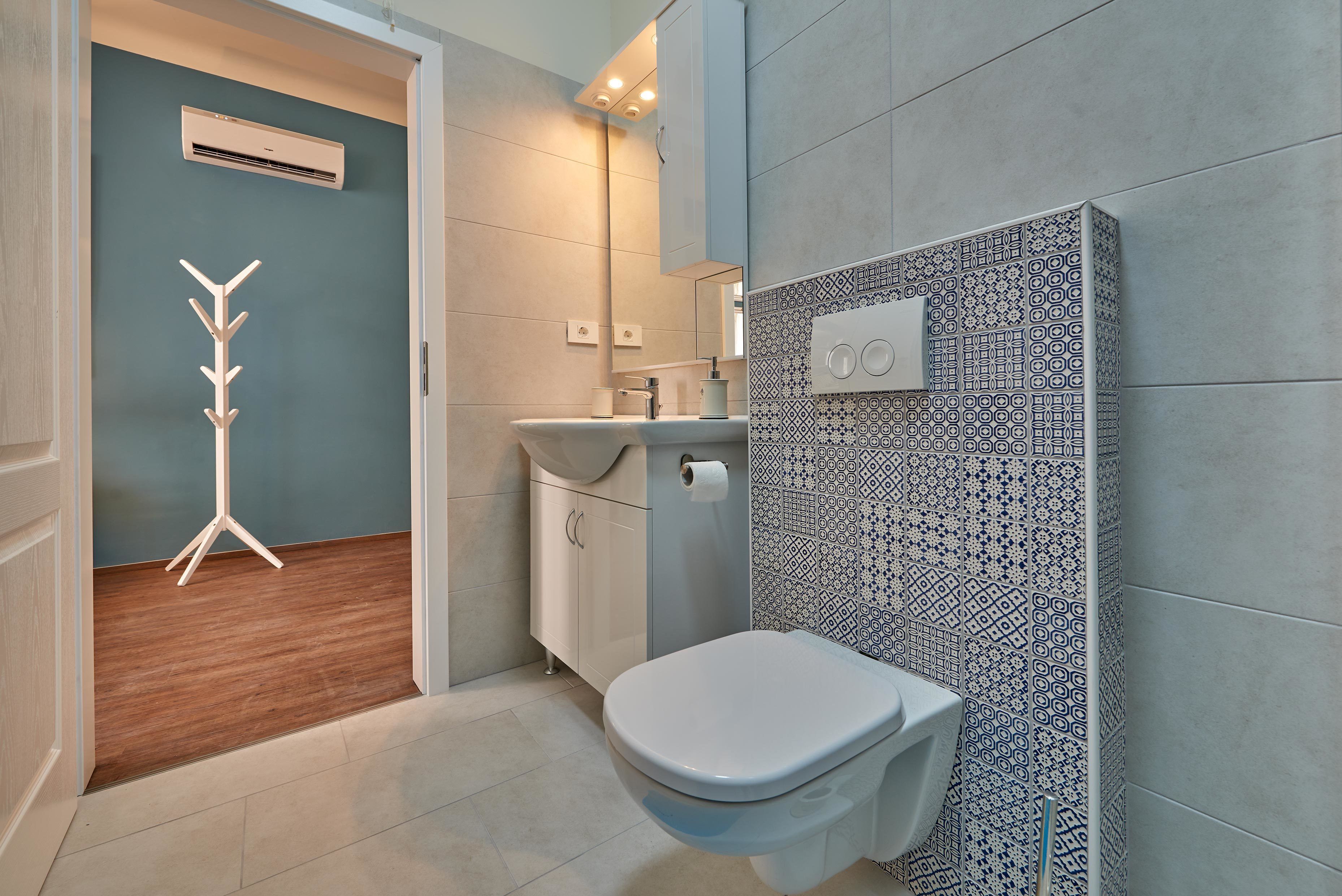 A modern bathroom with a white toilet, a wooden coat rack, a sink with a mirror, and patterned tiles on the walls.