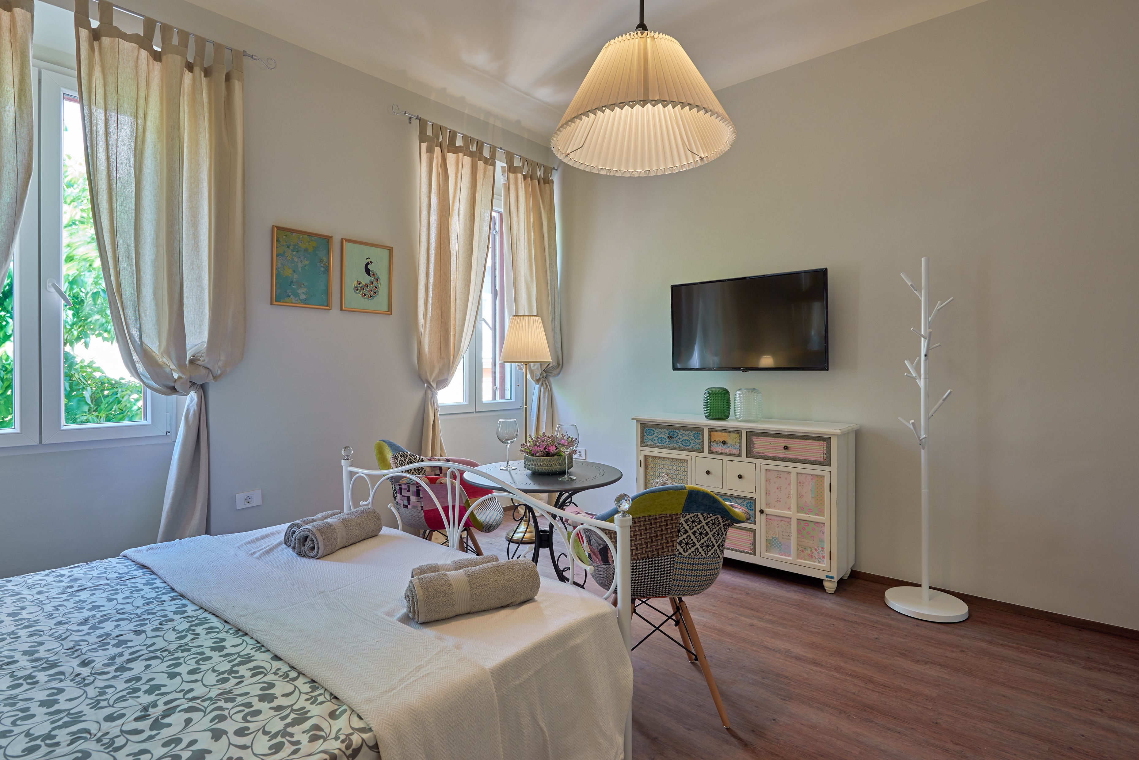 Quarto bem iluminado com cama, mesa de cabeceira, cadeira e televisão na parede.