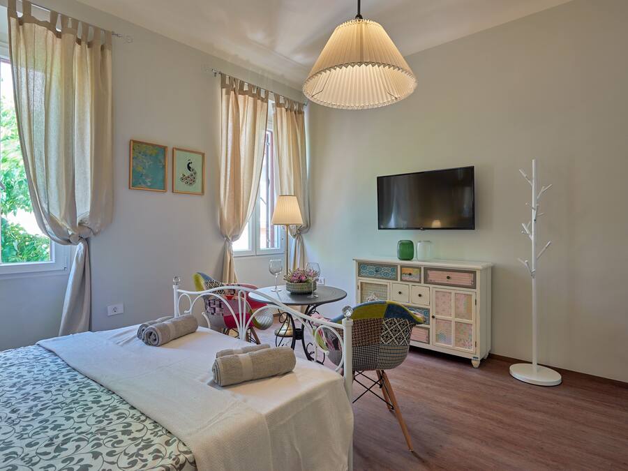 Quarto bem iluminado com cama, mesa de cabeceira, cadeira e televisão na parede.