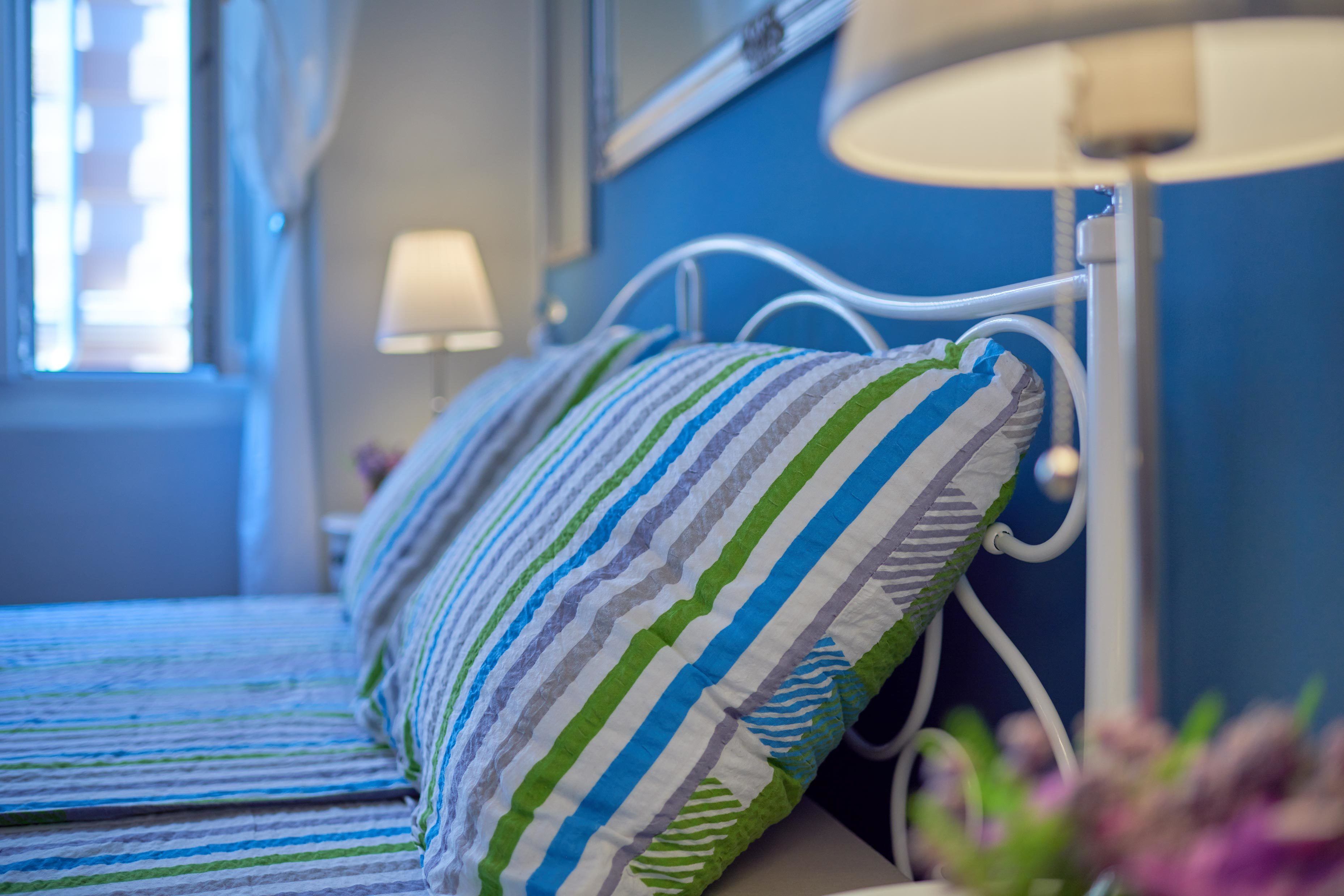 Una cama con almohadas a rayas y un cabecero de metal blanco en una habitación con paredes azules y una ventana.