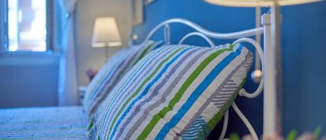 Una cama con almohadas a rayas y un cabecero de metal blanco en una habitación con paredes azules y una ventana.