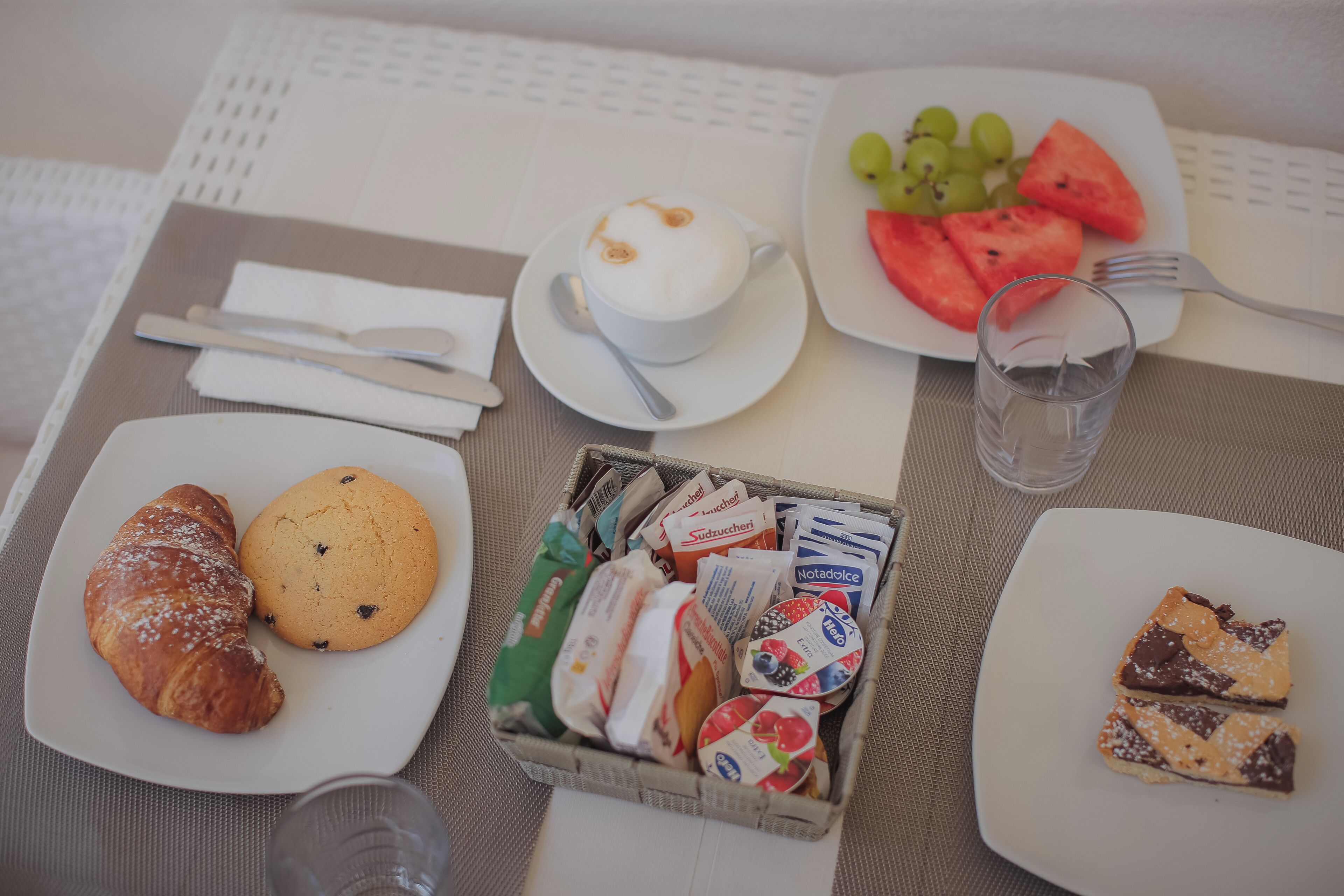 A breakfast setting with croissants, cookies, a cup of coffee, a plate of watermelon and grapes, a glass of water, and a basket of assorted pastries.