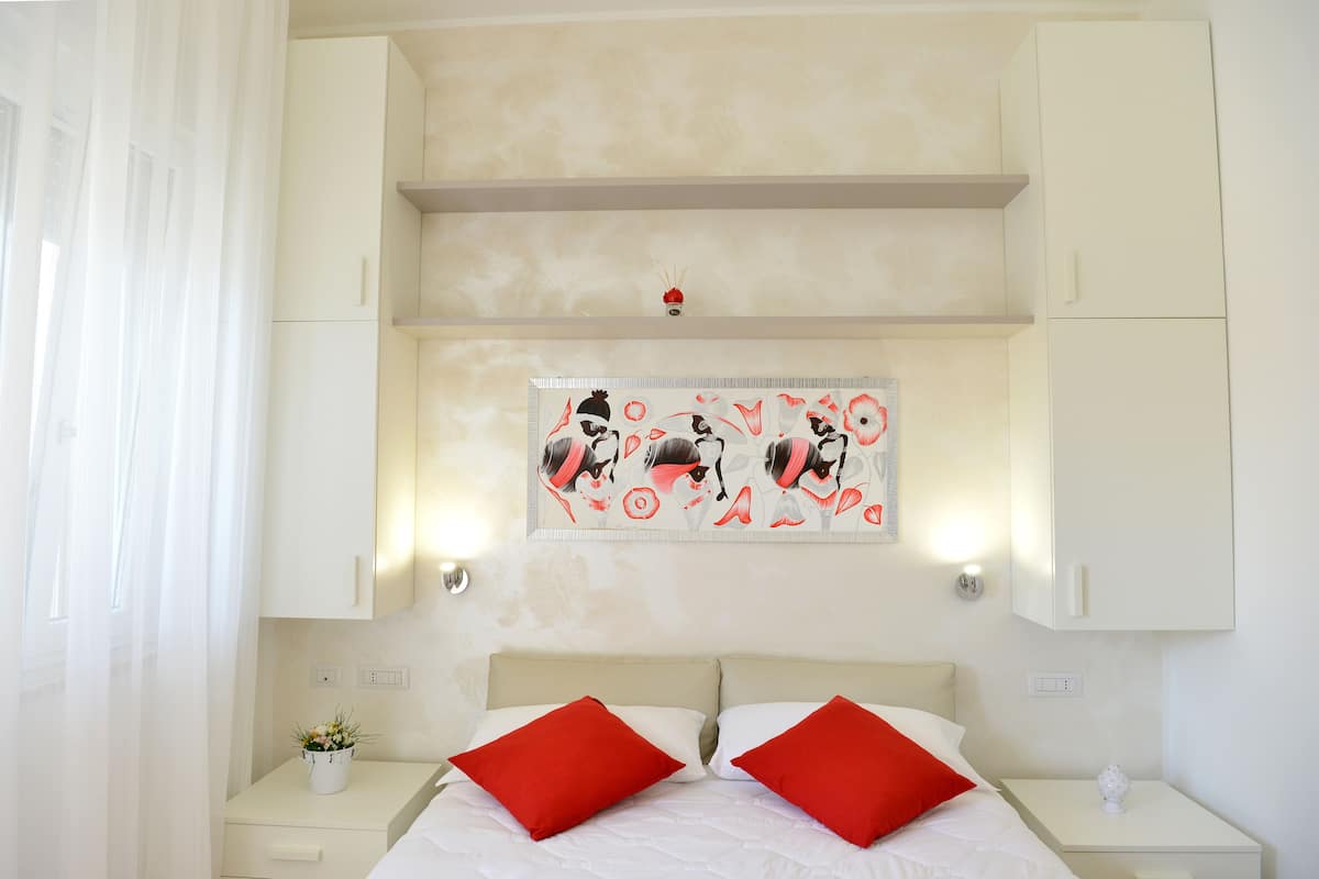 A bedroom with a bed, red pillows, a nightstand with a plant, and a wall-mounted artwork.