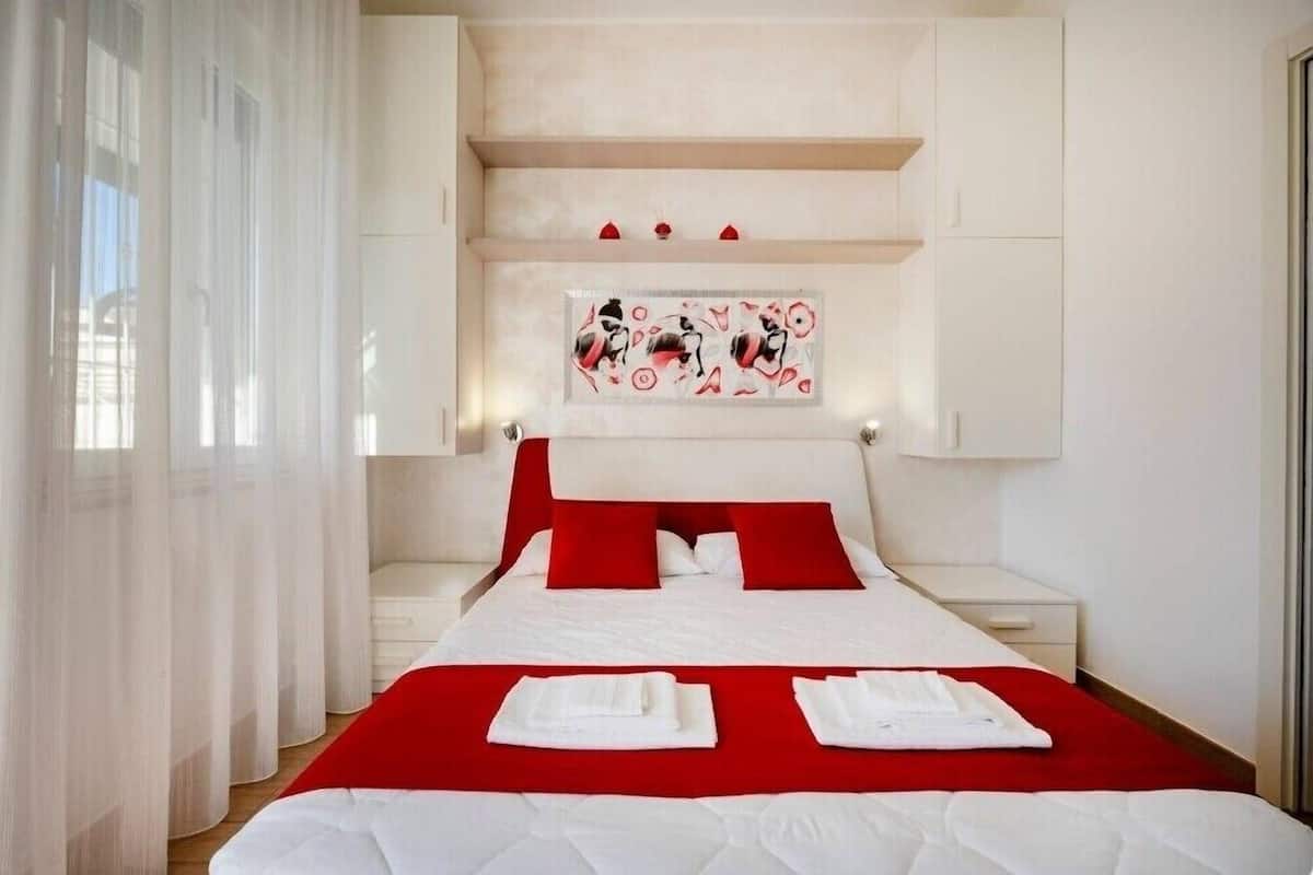 A hotel room with a bed, white curtains, a painting on the wall, and a red accent pillow.